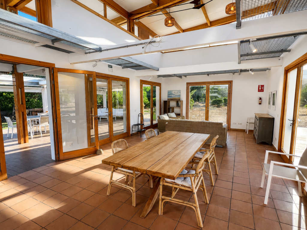 Amplio comedor con mesa de madera, zona de estar, vigas vistas y grandes ventanales abiertos hacia los jardines de la finca.